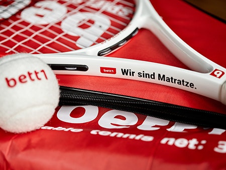 Foto: Im Fokus der Hals eines Tennisschlägers mit der Aufschrift „bett1 – Wir sind Matratze.“; unscharf davor liegt weißer bett1-Tennisball.