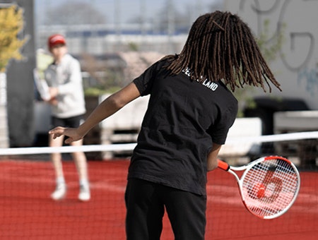 Foto: Ein Kind spielt einen roten Tennisball über das Netz zu einem anderen Kind.
